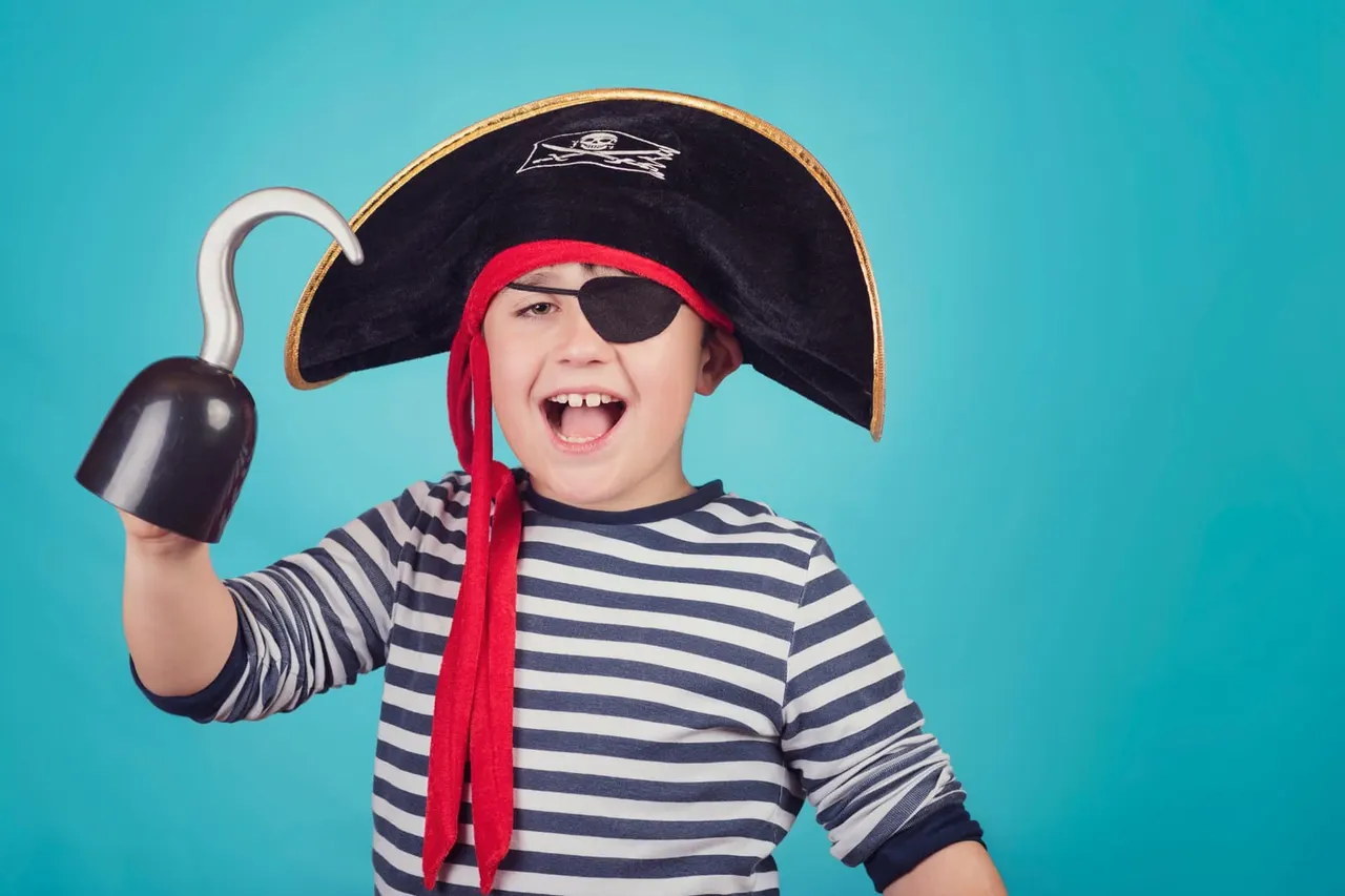 Child in pirate costume with tricorn hat, eye patch and striped shirt holding a toy bomb against a turquoise background.