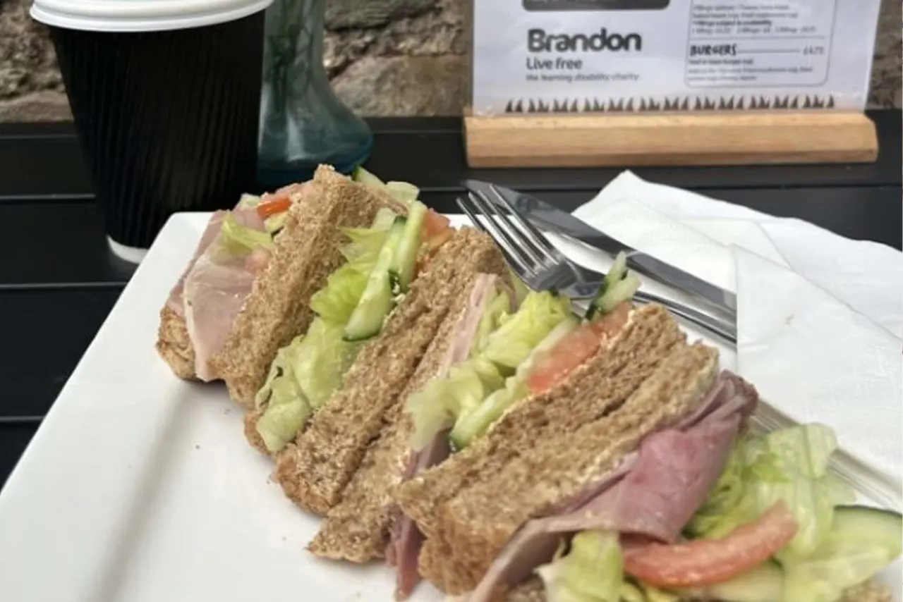 A club sandwich with wholemeal bread, lettuce, ham and tomato, served on a white plate with a fork, alongside a black coffee cup and menu.
