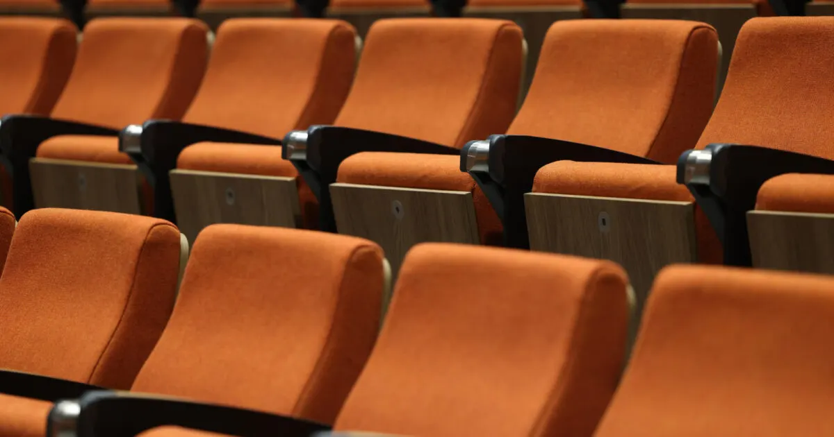 Rows of empty orange upholstered theatre seats with black armrests and wooden side panels.