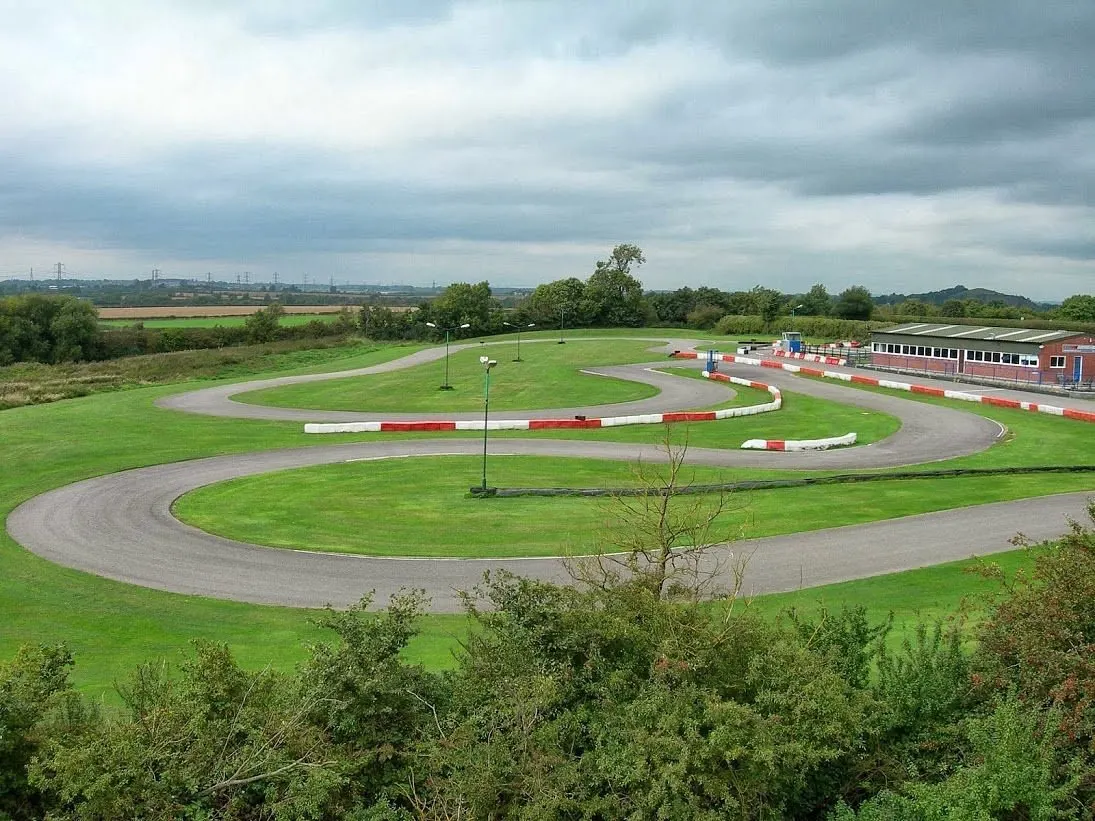 Go-kart racing track with red and white barriers, green grass infield, and brick buildings beside rural farmland.