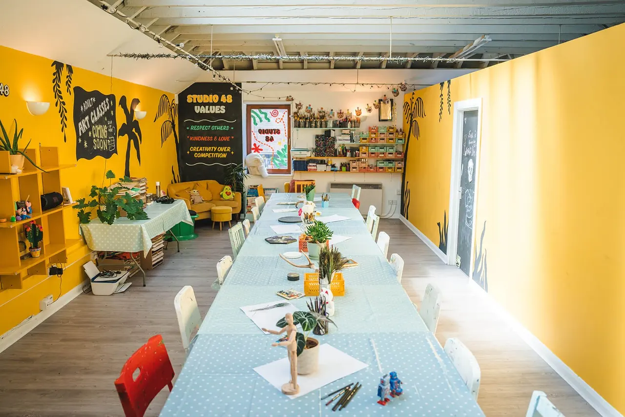 A bright yellow art studio with long tables set up for creative activities, plants, art supplies, and display shelves of finished work.
