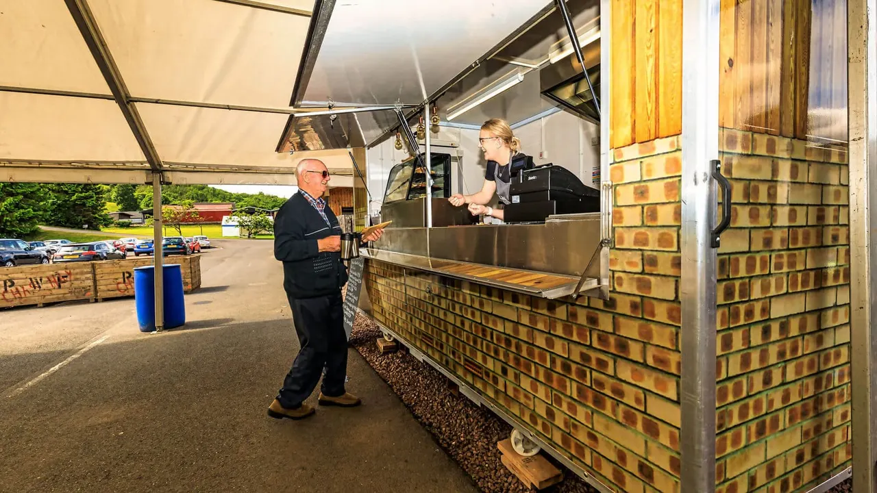 A man in glasses and dark clothing approaches a food service counter where a staff member serves him under a clear canopy, with parked cars visible in the background.