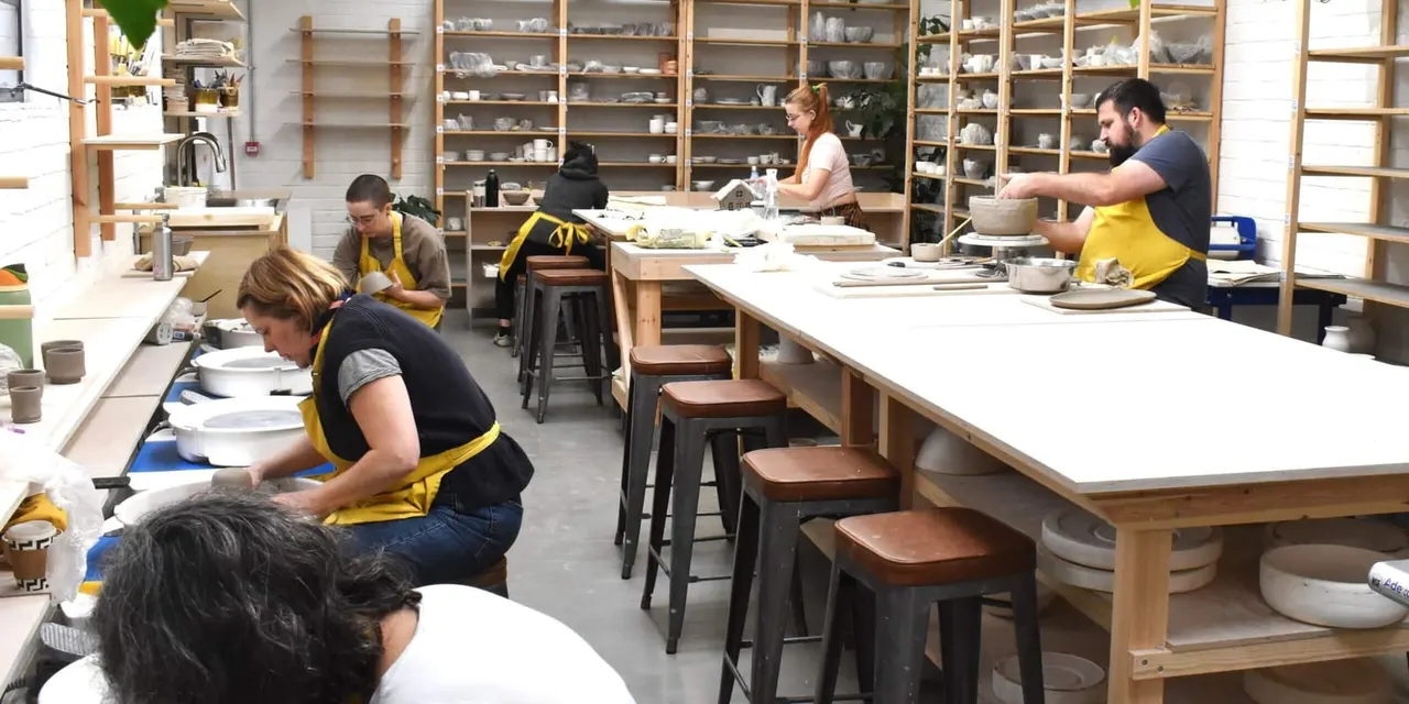 Multiple people in yellow aprons working at pottery wheels and tables in a ceramic studio with shelving displaying finished pottery pieces.