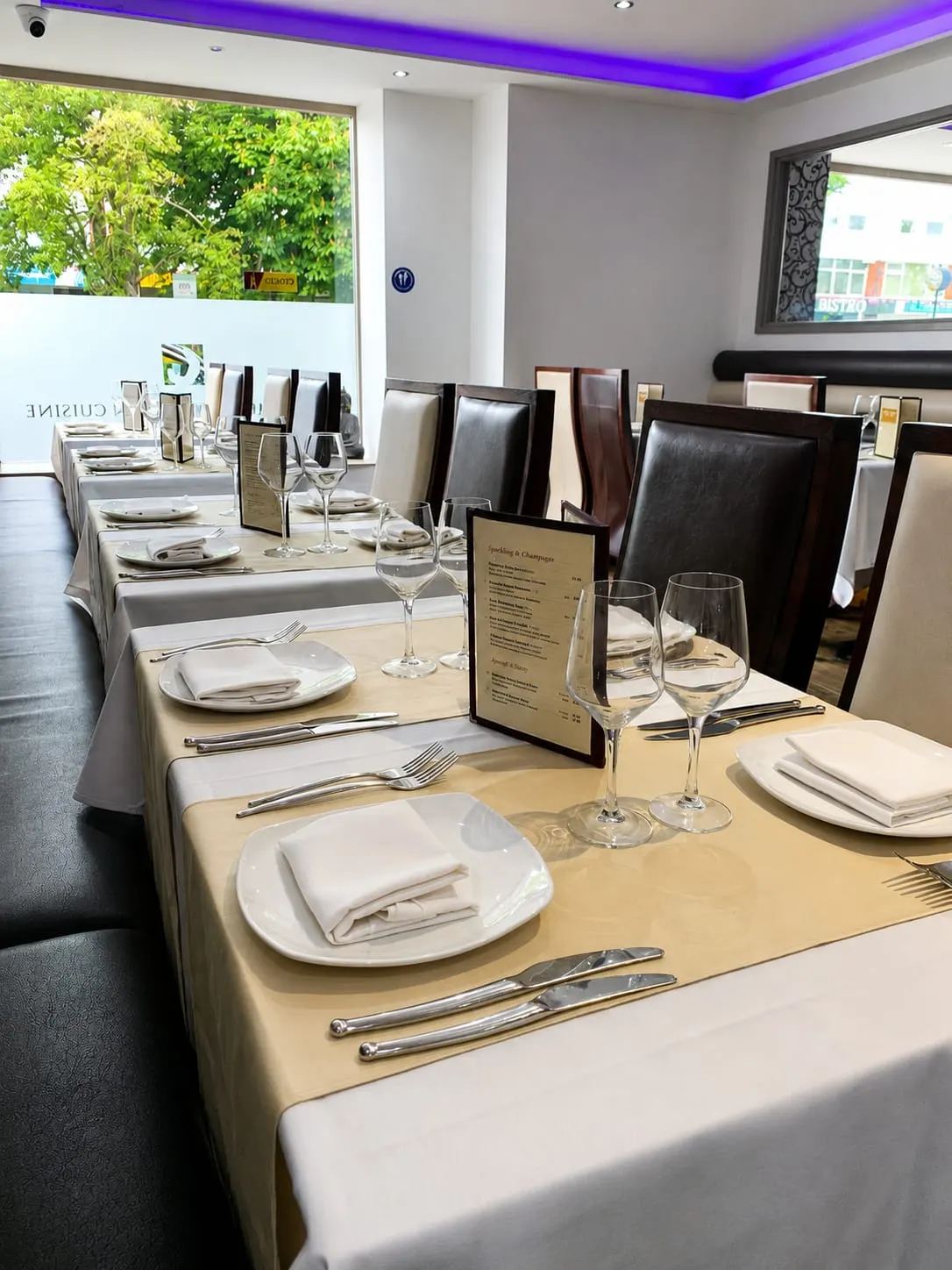 A restaurant dining room set with formal table settings, place plates, glasses, cutlery and napkins along a beige table with black chairs.