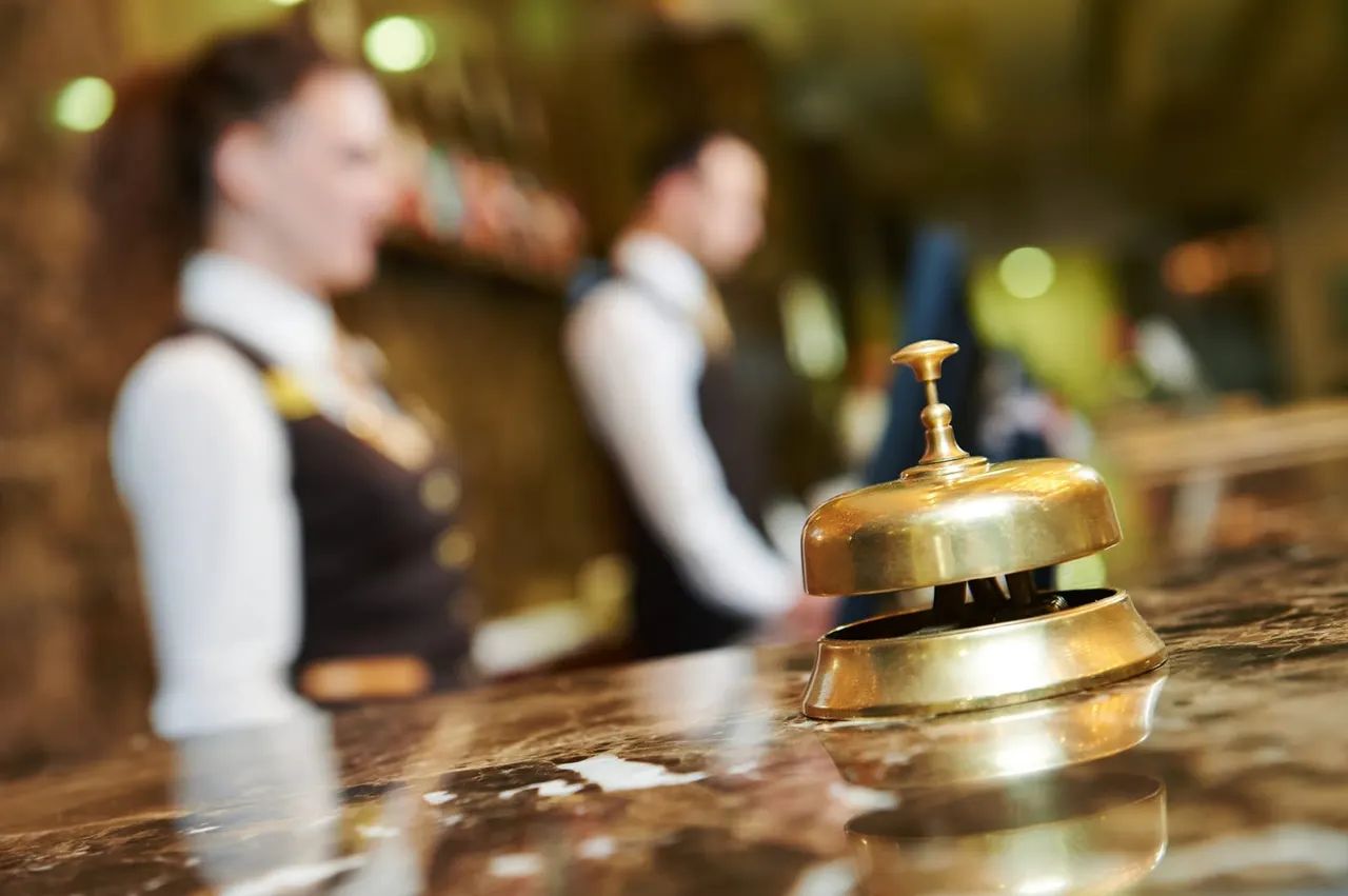 A brass reception bell on a wooden counter with hotel staff in uniform blurred in the background.