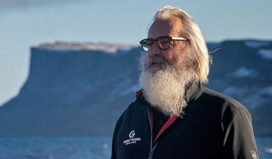 Man with long white beard and glasses stands outdoors with coastal cliffs in the background.