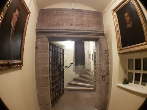 Stone passageway with large wooden door and Latin inscribed archway, flanked by framed portraits on cream walls.
