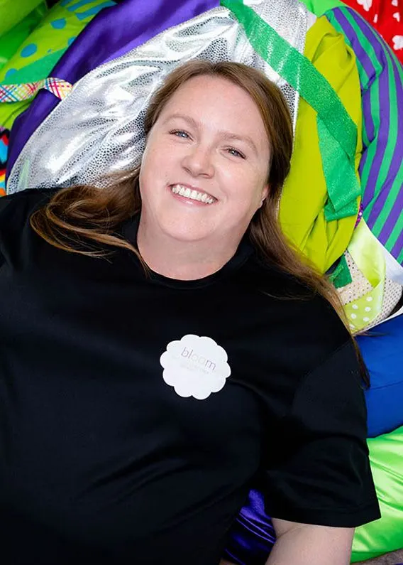 Woman smiling at camera surrounded by colourful play equipment and soft fabrics.
