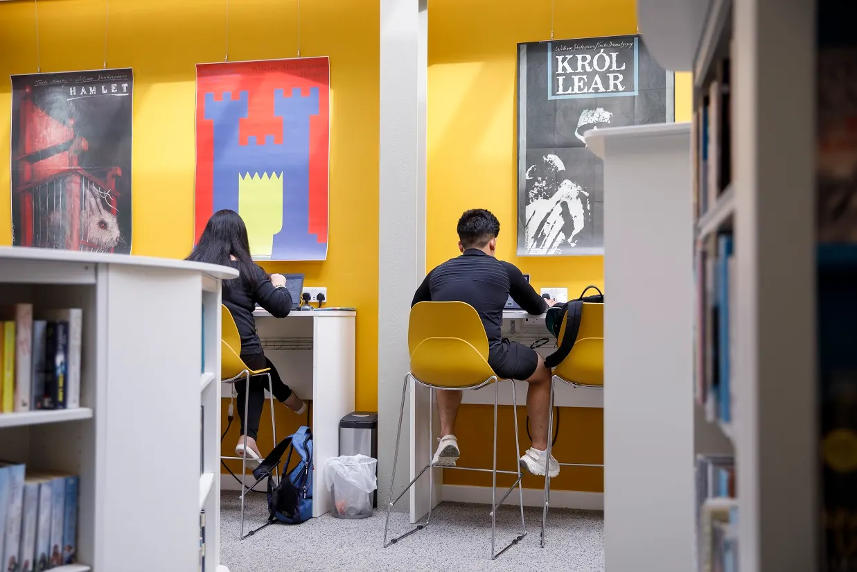 Two people seated at individual desks in a bright yellow study area with Shakespeare posters on the wall.