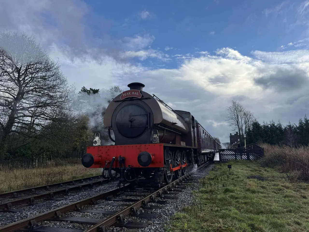 Vintage steam locomotive with red base and attached carriages on railway track, surrounded by bare trees and countryside.