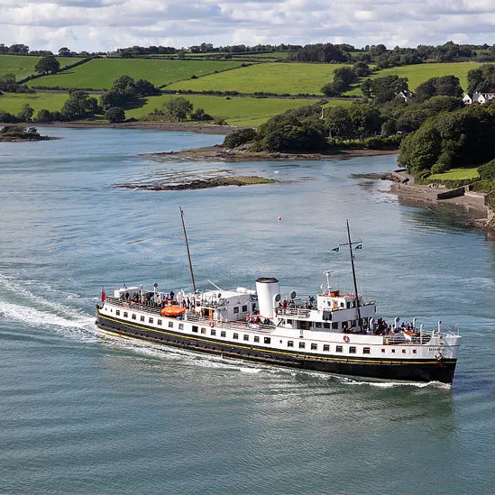 Passenger ship steaming through blue-green waters with green fields and woodland coastline behind.