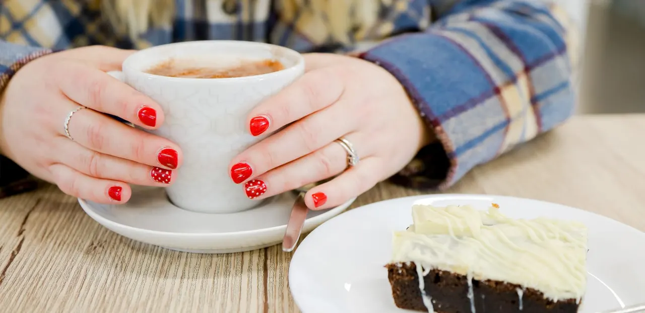 Person with red nail varnish holding a cup of coffee next to a slice of cake on a wooden table.