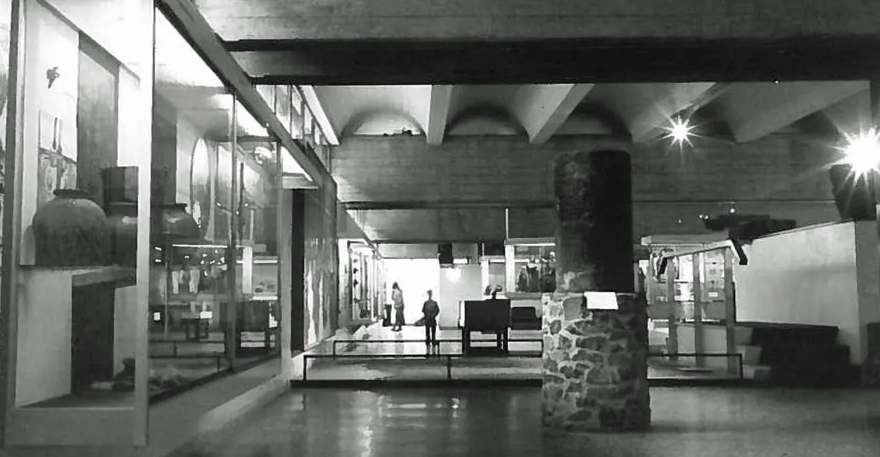 Interior of museum gallery with concrete brutalist architecture, display cases, and a few visitors walking through the space.