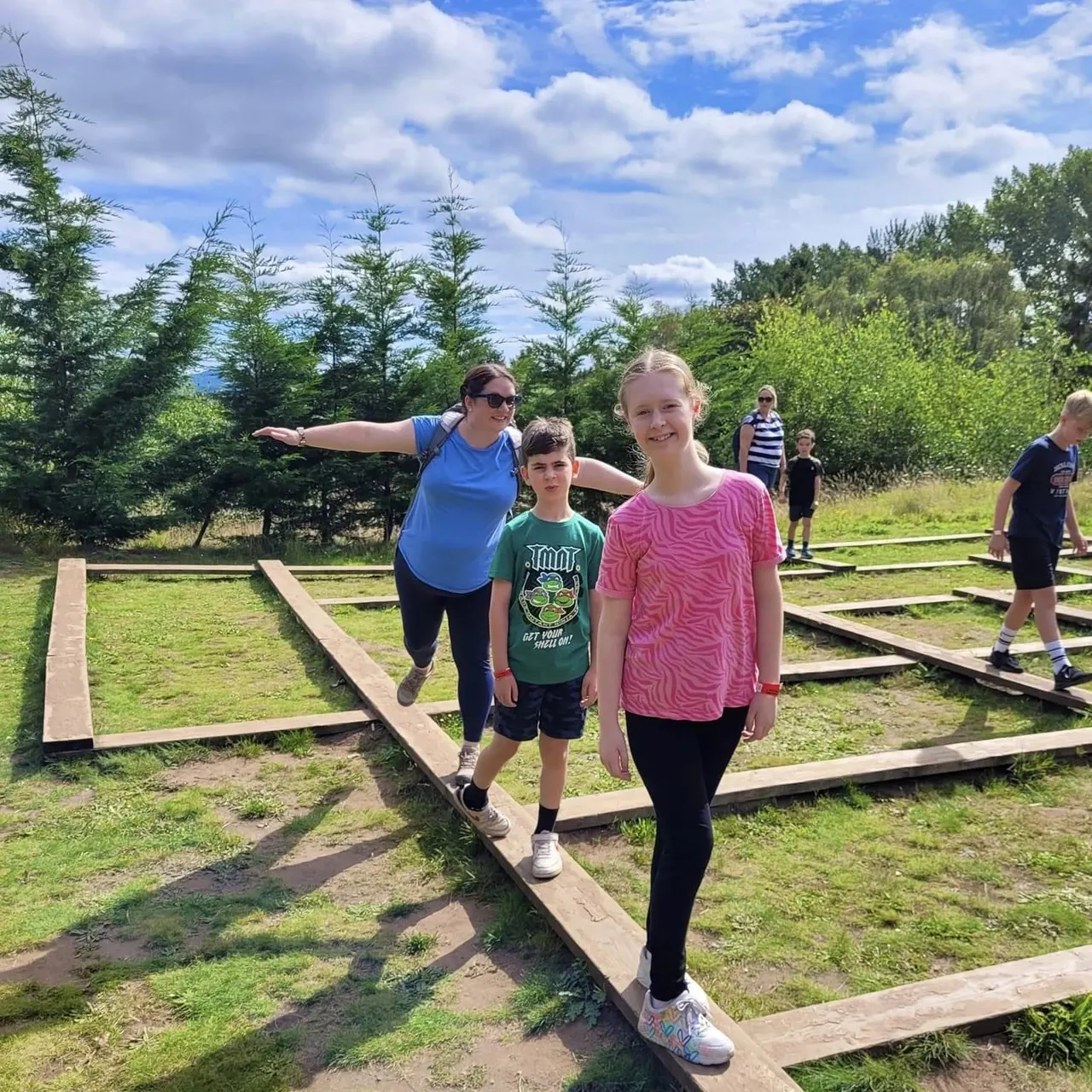 Adults and children balancing on wooden beams during an outdoor activity course among trees.