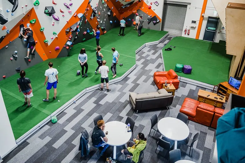 Overhead view of indoor climbing centre with visitors on green floor and colourful wall-mounted climbing holds.