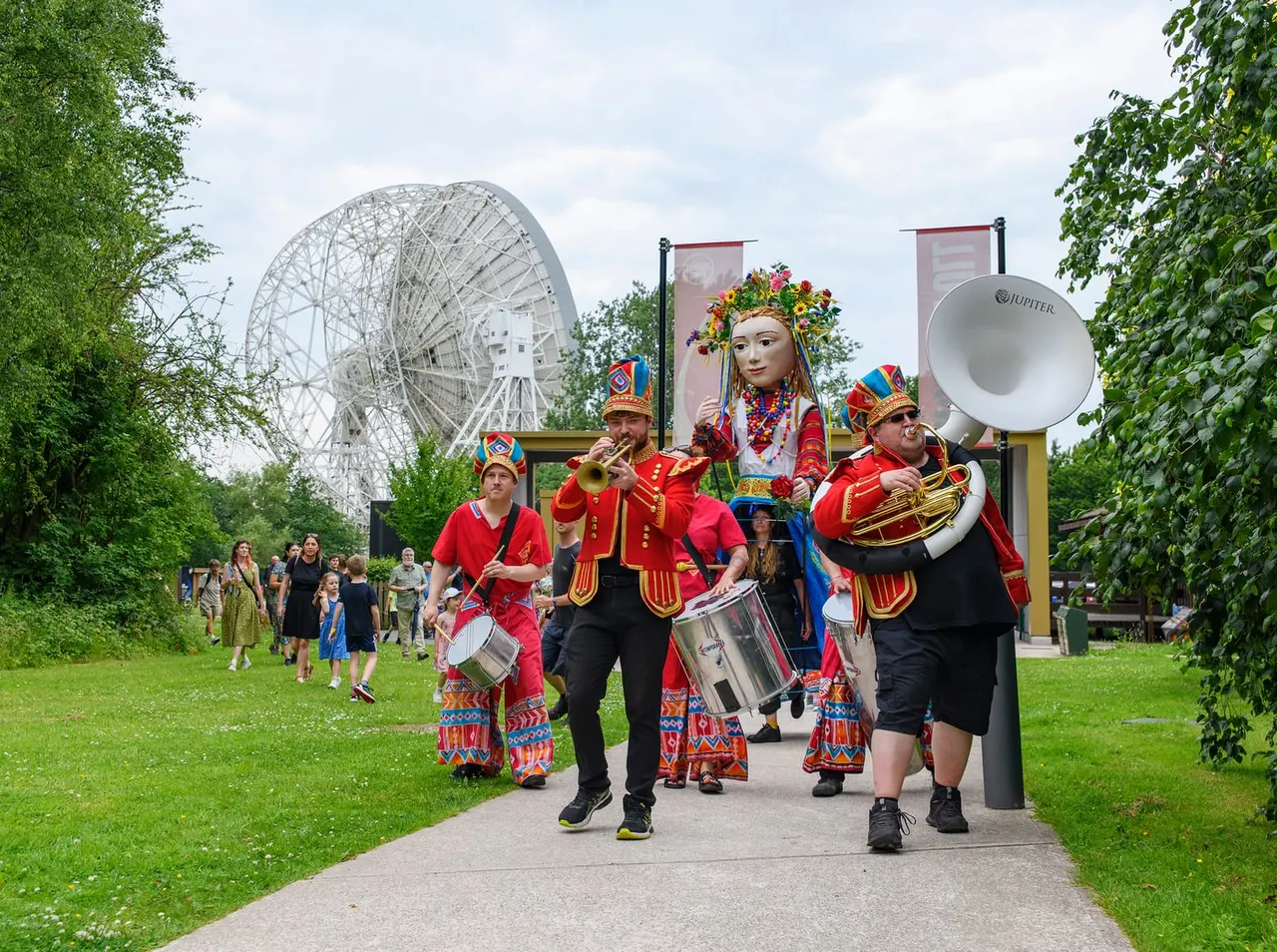 Performers in colourful traditional costume parade with brass instruments past visitors on a grass path, with a large radio telescope in the background.