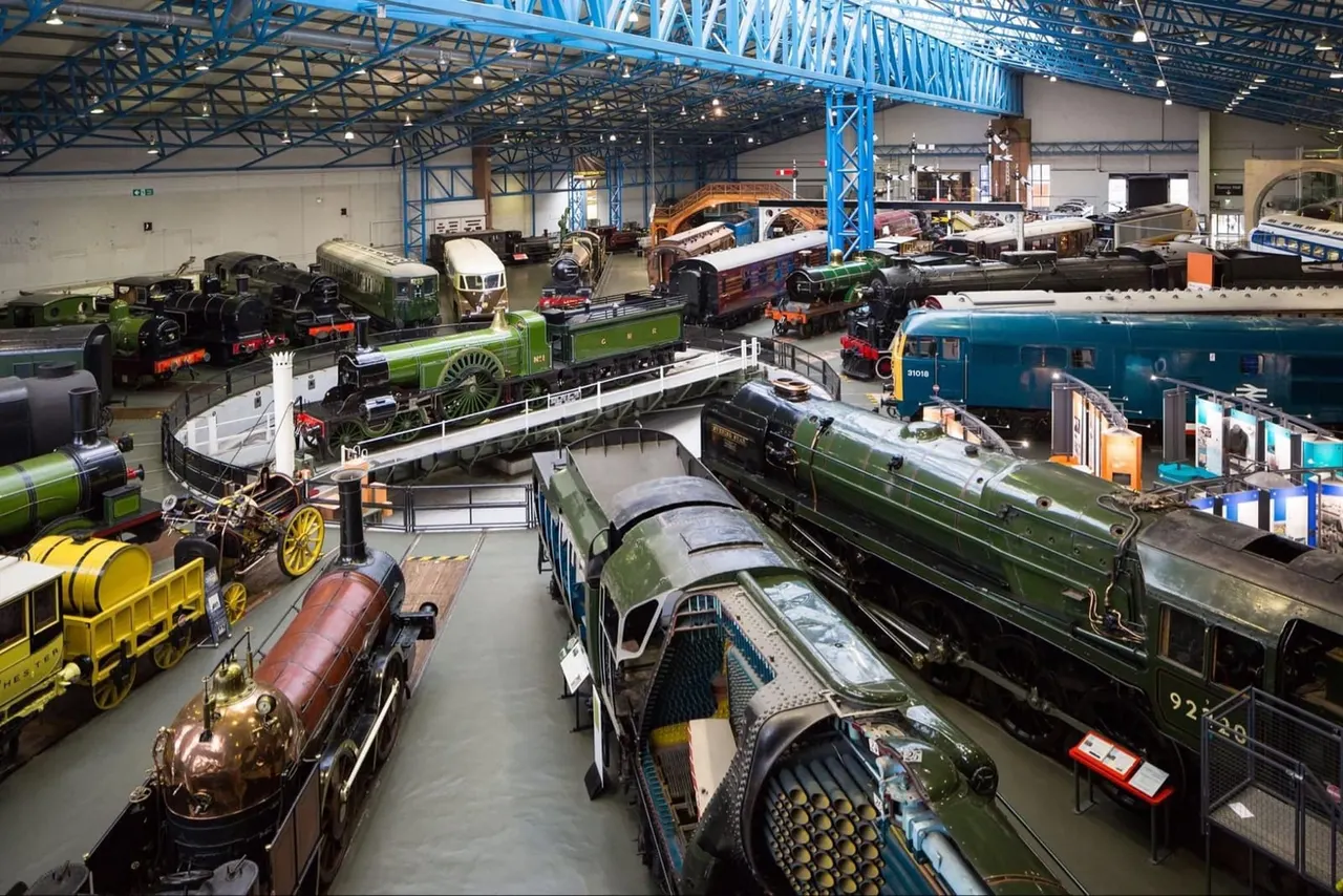 Historic steam locomotives and railway carriages displayed in a large industrial museum hall with blue steel framework.