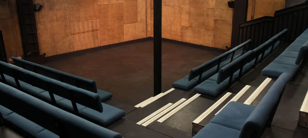 Empty theatre seating area with black bench seats facing a dark wooden floor and terracotta-tiled walls.