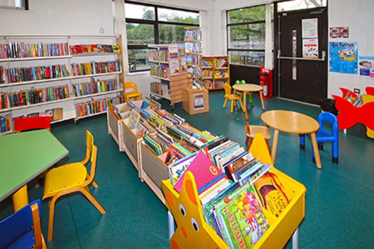 Bright children's library area with shelves of books, colourful chairs and tables, and display racks of children's reading materials.