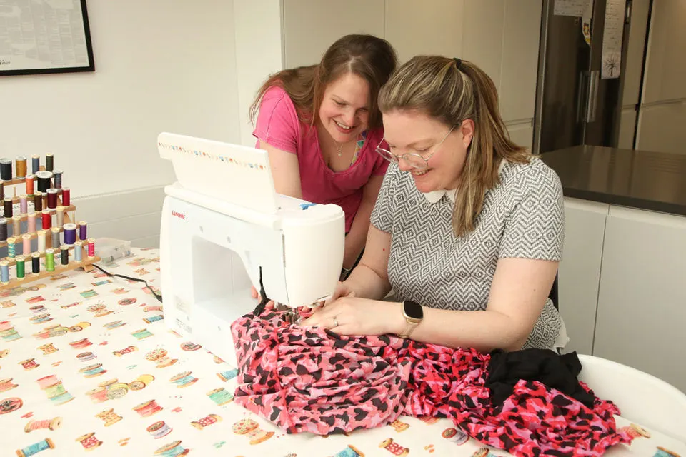 Two women using a sewing machine to work with pink patterned fabric at a craft table.