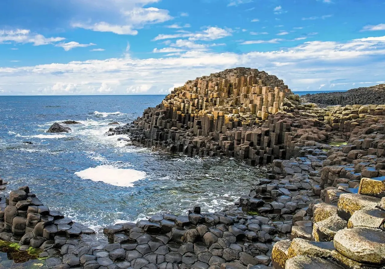 Hexagonal basalt rock formations rise from the sea along a rocky coastline under blue sky with white clouds.