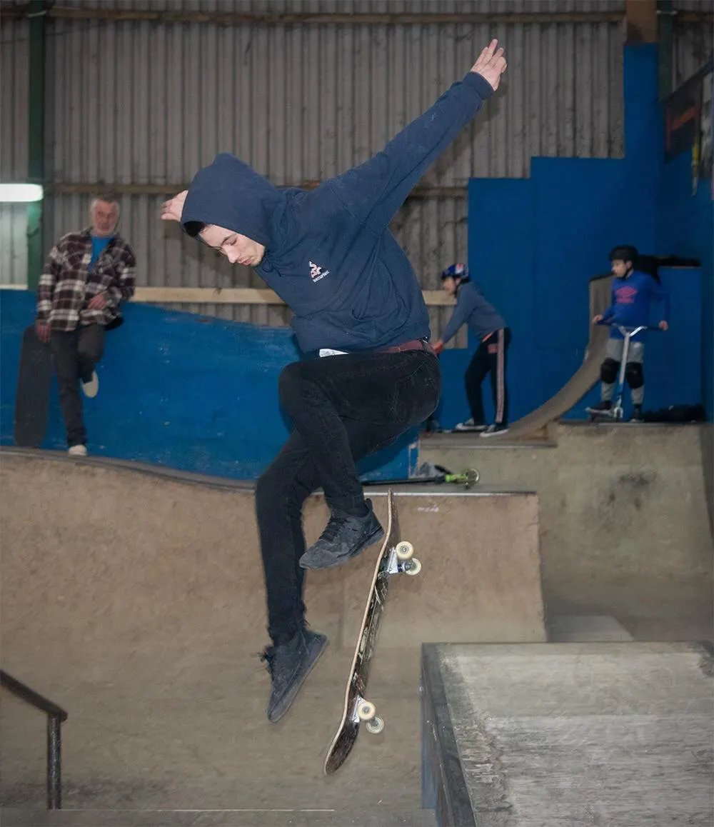 Young skateboarder performing a trick mid-air above a concrete ramp, with spectators watching at an indoor skate park.