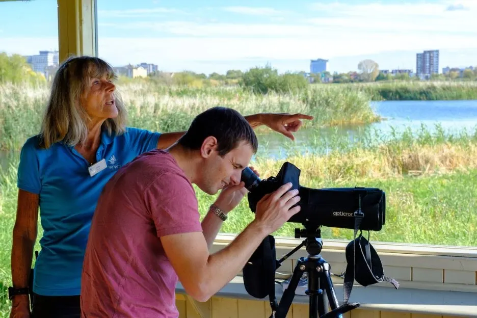 A staff member in blue points out features of the wetland landscape while a visitor peers through a spotting scope at reeds, water and distant buildings.
