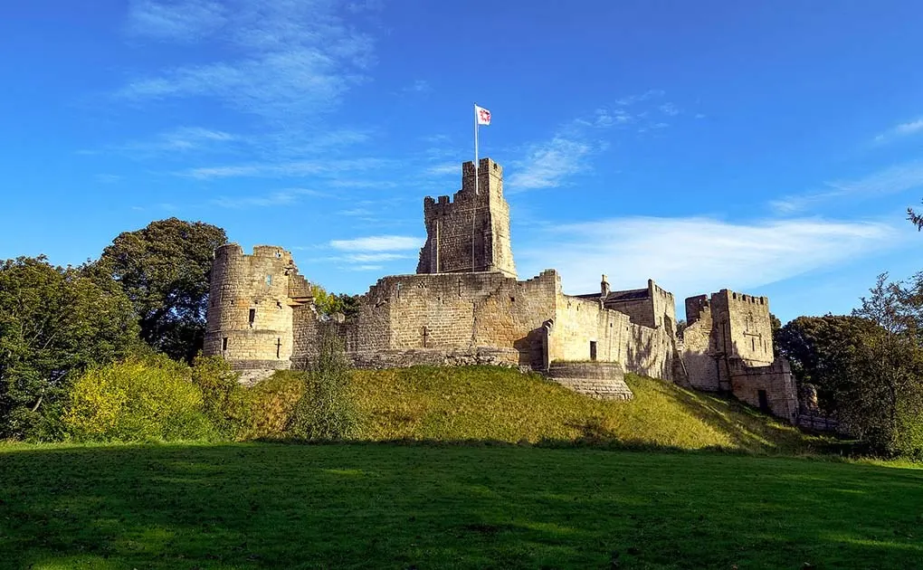 Medieval stone castle with central tower and flag, set on grassy mound surrounded by trees under blue sky.