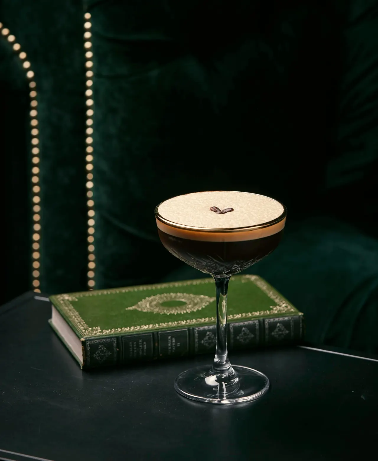 Cocktail with foam top and garnish displayed beside a green leather-bound book against dark green backdrop with string lights.
