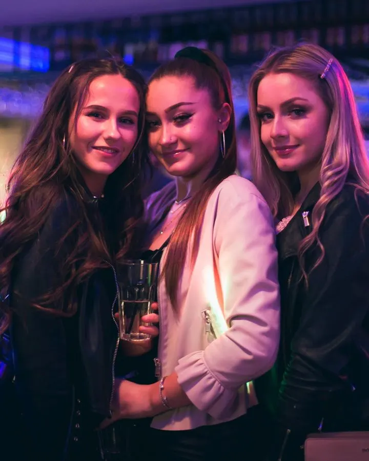Three young women posing together at a nightclub with purple and blue lighting, holding drinks.