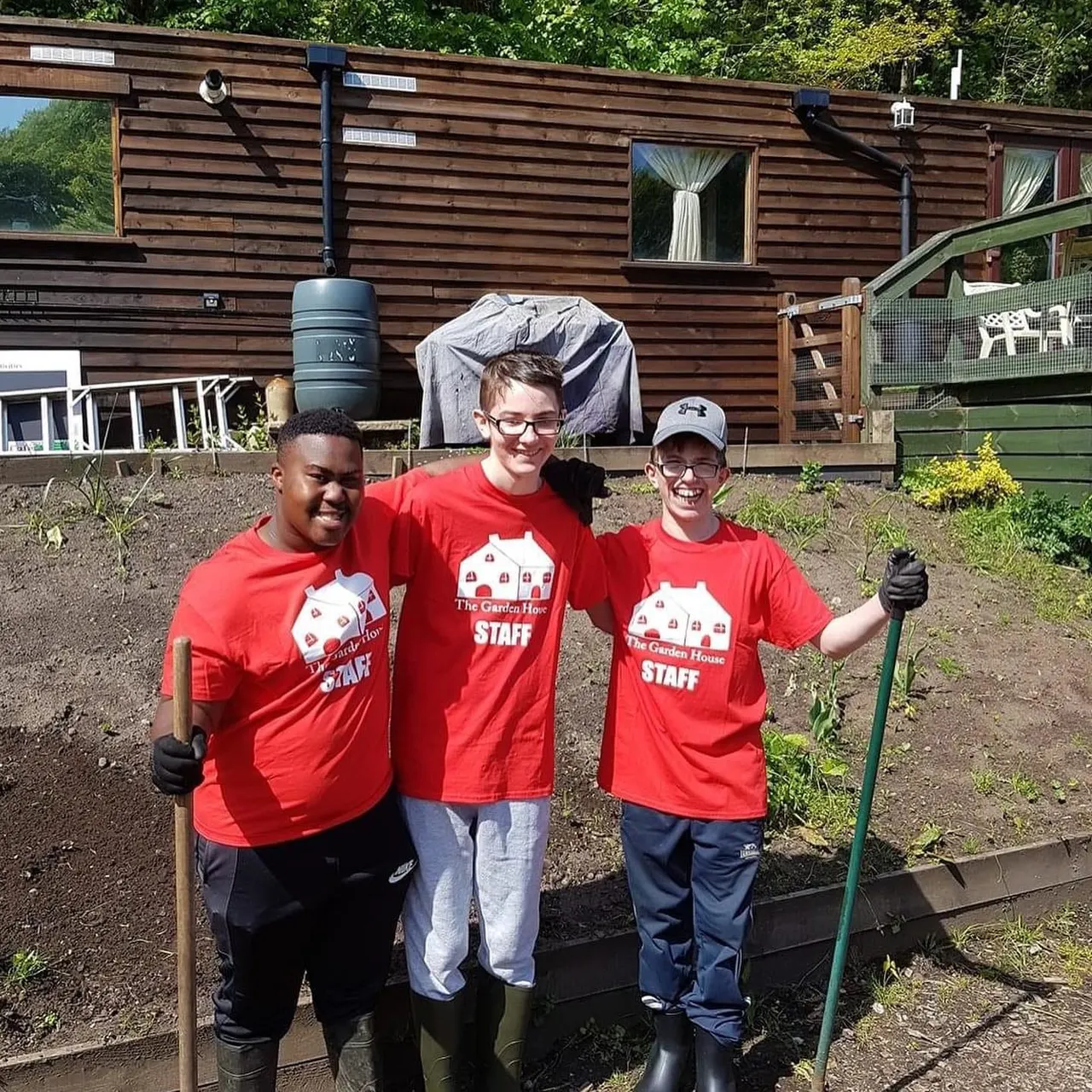 Three smiling staff members in red Garden House t-shirts holding gardening tools in a vegetable garden plot beside a wooden lodge building.