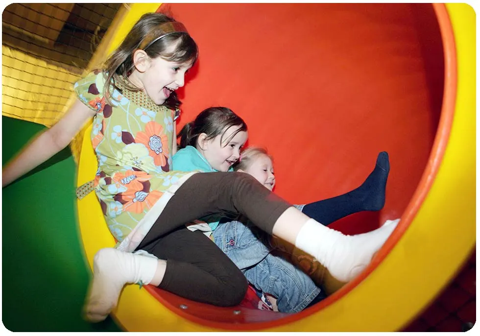 Three young children sliding down a brightly coloured tube slide in an indoor soft play area.