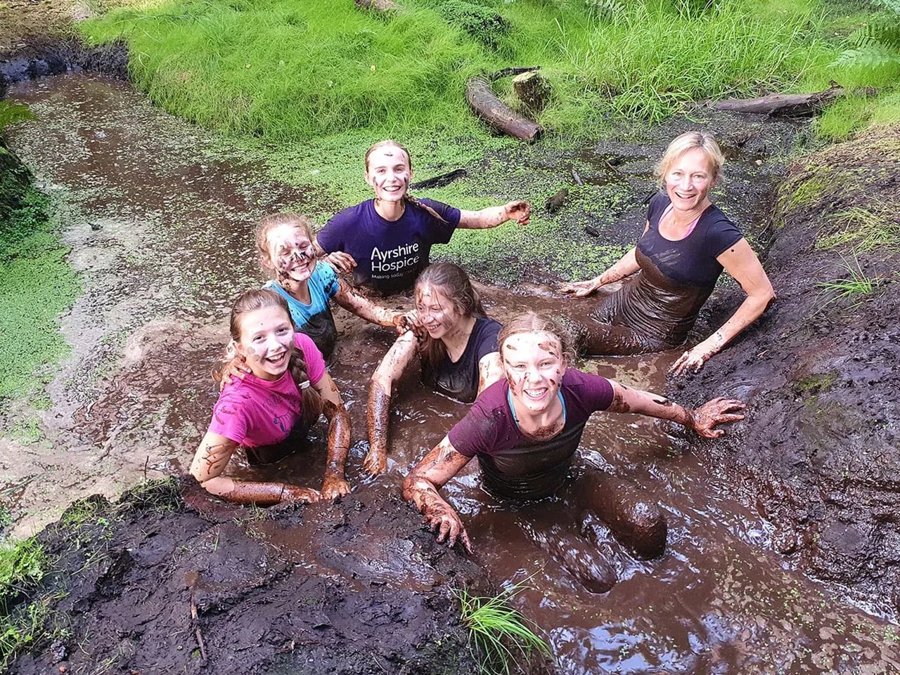Adults and children enjoying a muddy outdoor activity in a natural stream surrounded by grass and fallen logs.