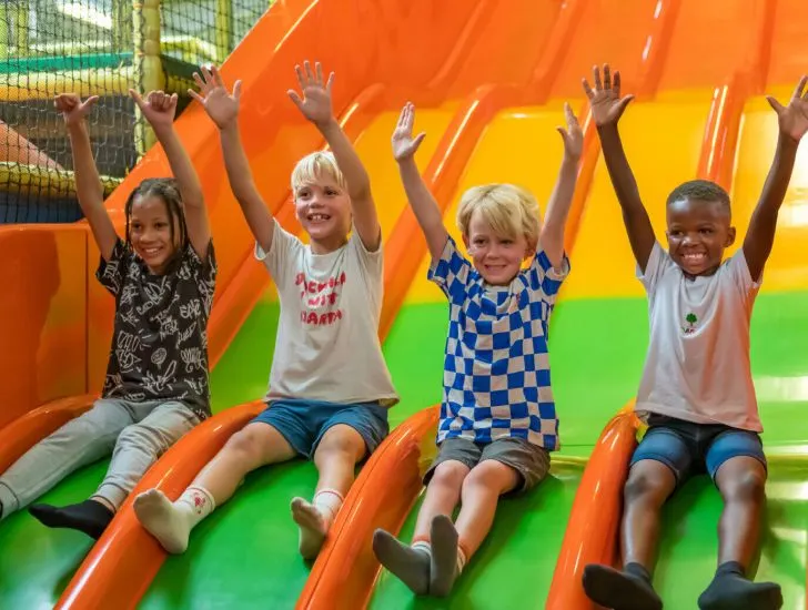 Four children sitting on a colourful inflatable slide with their arms raised in the air.