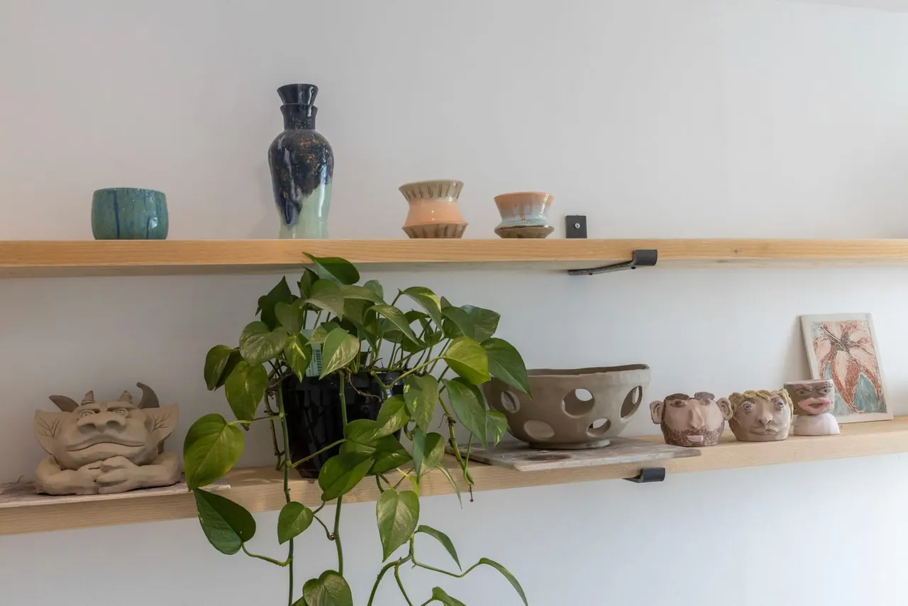Wooden wall shelves displaying pottery vessels, a potted plant, ceramic sculptural heads, and decorative artwork.