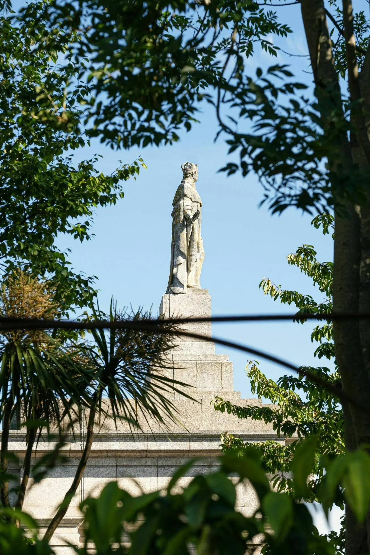 Stone statue of a robed figure on a tall plinth, viewed through trees and foliage with clear sky behind.
