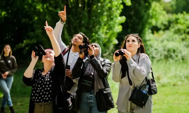 Group of visitors holding binoculars and looking upward in a tree-lined park setting.