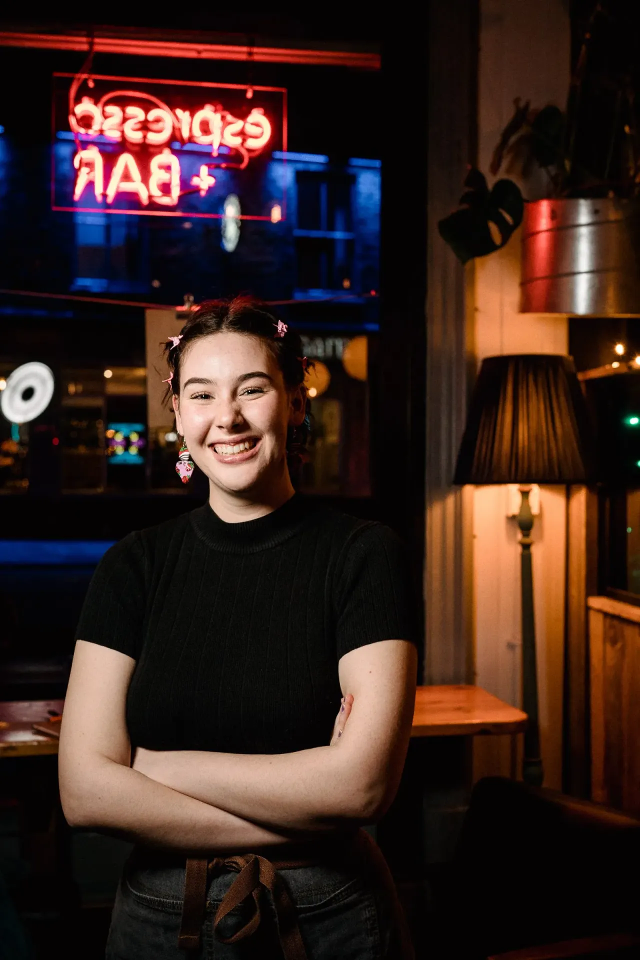 Young woman smiling at camera inside a bar or social venue with neon signage and decorative lighting behind her.