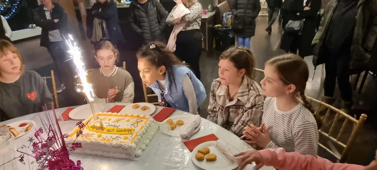Children gathered around a decorated birthday cake with sparklers whilst adults watch at an indoor celebration event.