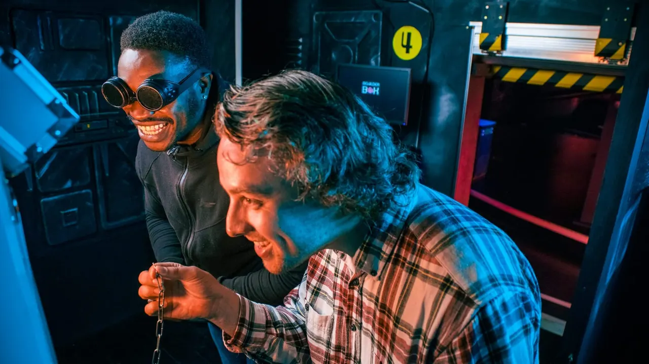 Two visitors playing an escape room game in a dark industrial-themed space lit with blue and red lighting.