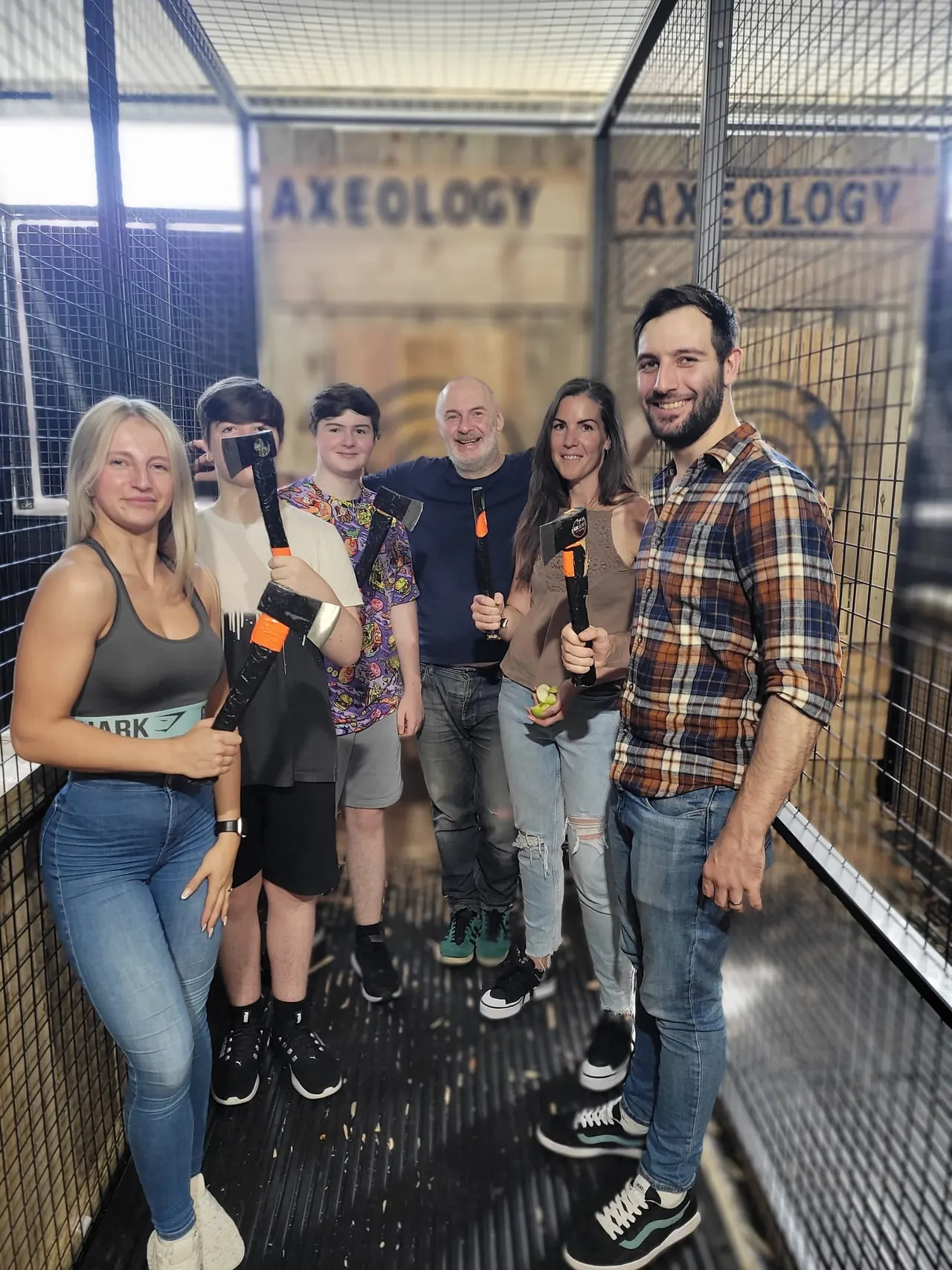 Group of six people holding axes inside an axe-throwing bay with netting and signage.