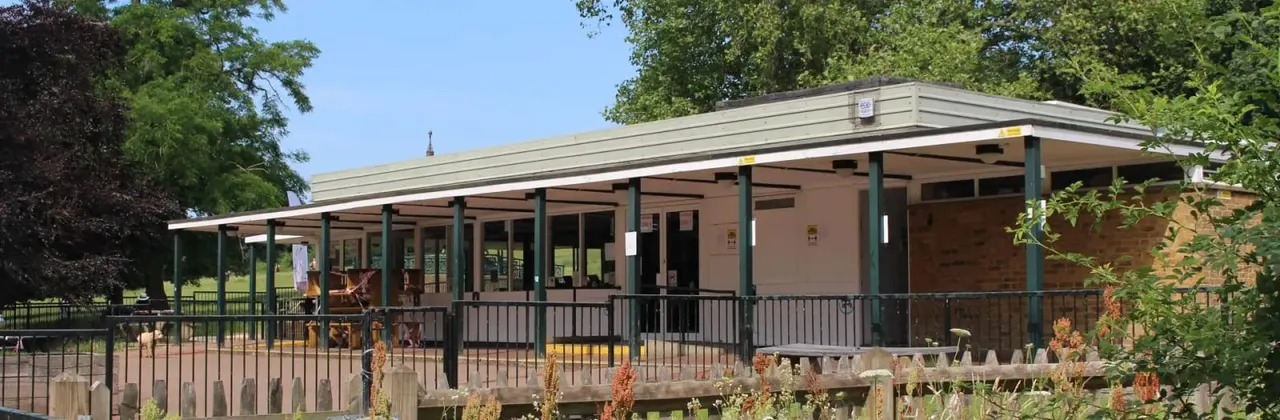 Modern pavilion building with covered verandah and railing, surrounded by trees and open green space on a sunny day.
