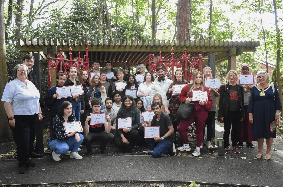 Large group of people of various ages posing together outdoors holding certificates under a wooden pergola with red decorative garland, surrounded by trees.