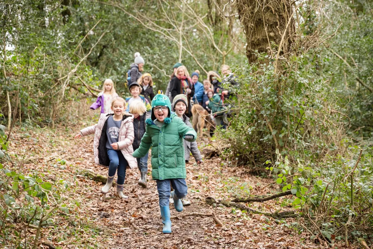 A group of children and adults walking and running together along a woodland trail surrounded by bare branches and green foliage.