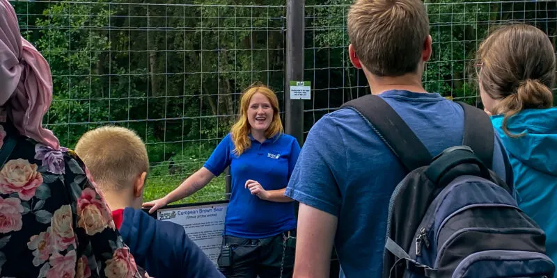 A staff member in a blue uniform engages with visitors of various ages at an animal enclosure behind a wire fence.