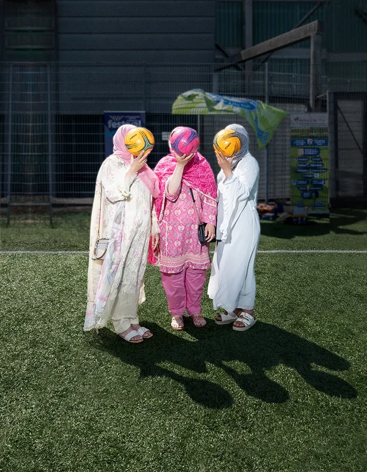 Three children in modest dress holding yellow and pink painted balls above their heads on astroturf pitch.