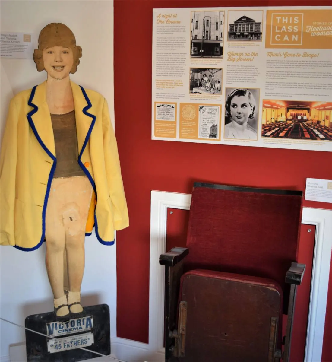 A museum display featuring a vintage cinema usherette mannequin in yellow jacket alongside period cinema seating and historical photographs and documents on a red wall.