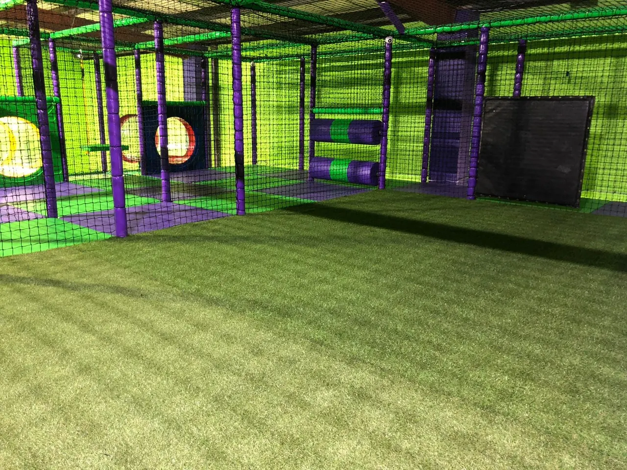 Indoor soft play arena with purple-framed netting enclosures, green mesh panels, and foam targets on artificial turf flooring.