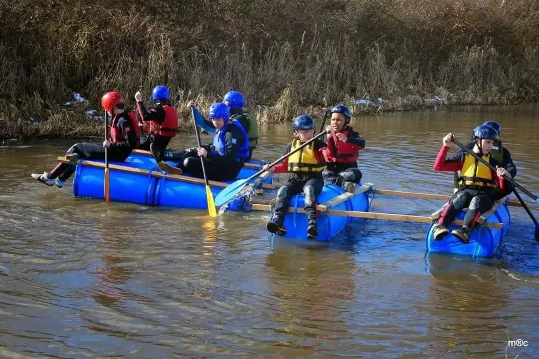 Groups of people in helmets and lifejackets paddling blue inflatable rafts on a shallow river.