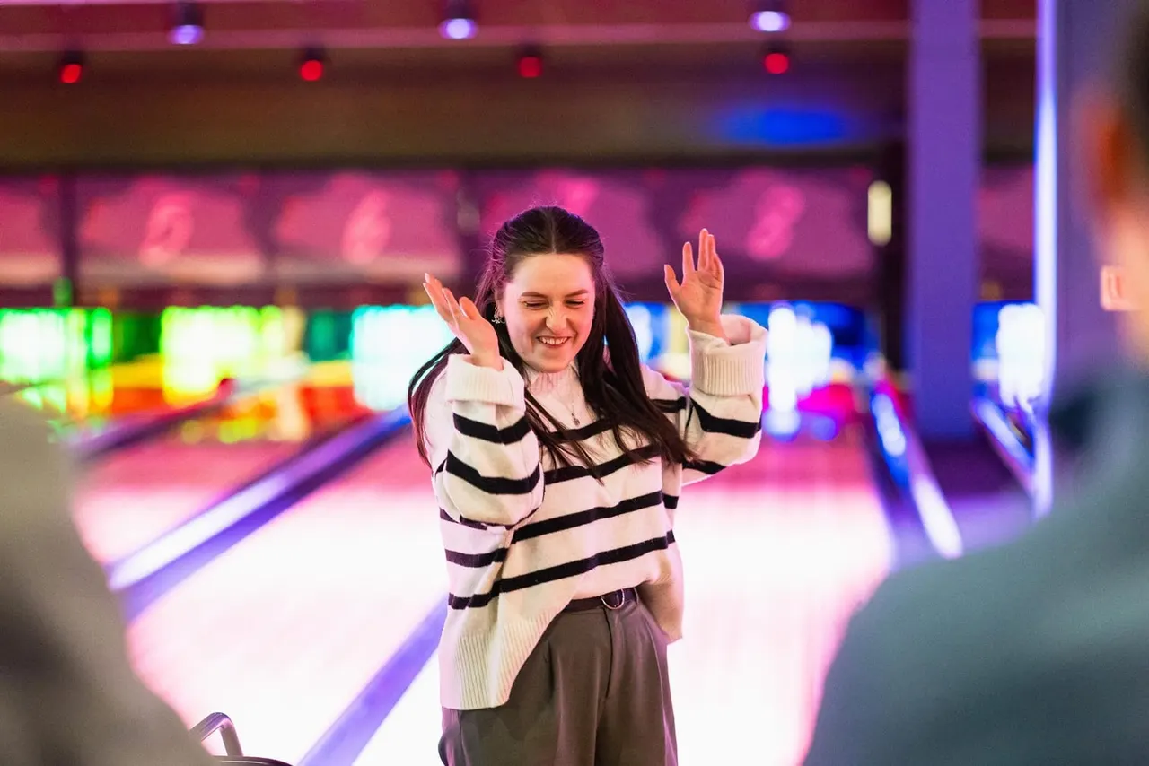 Young woman in striped sweater celebrating at a bowling alley with neon-lit lanes behind her.